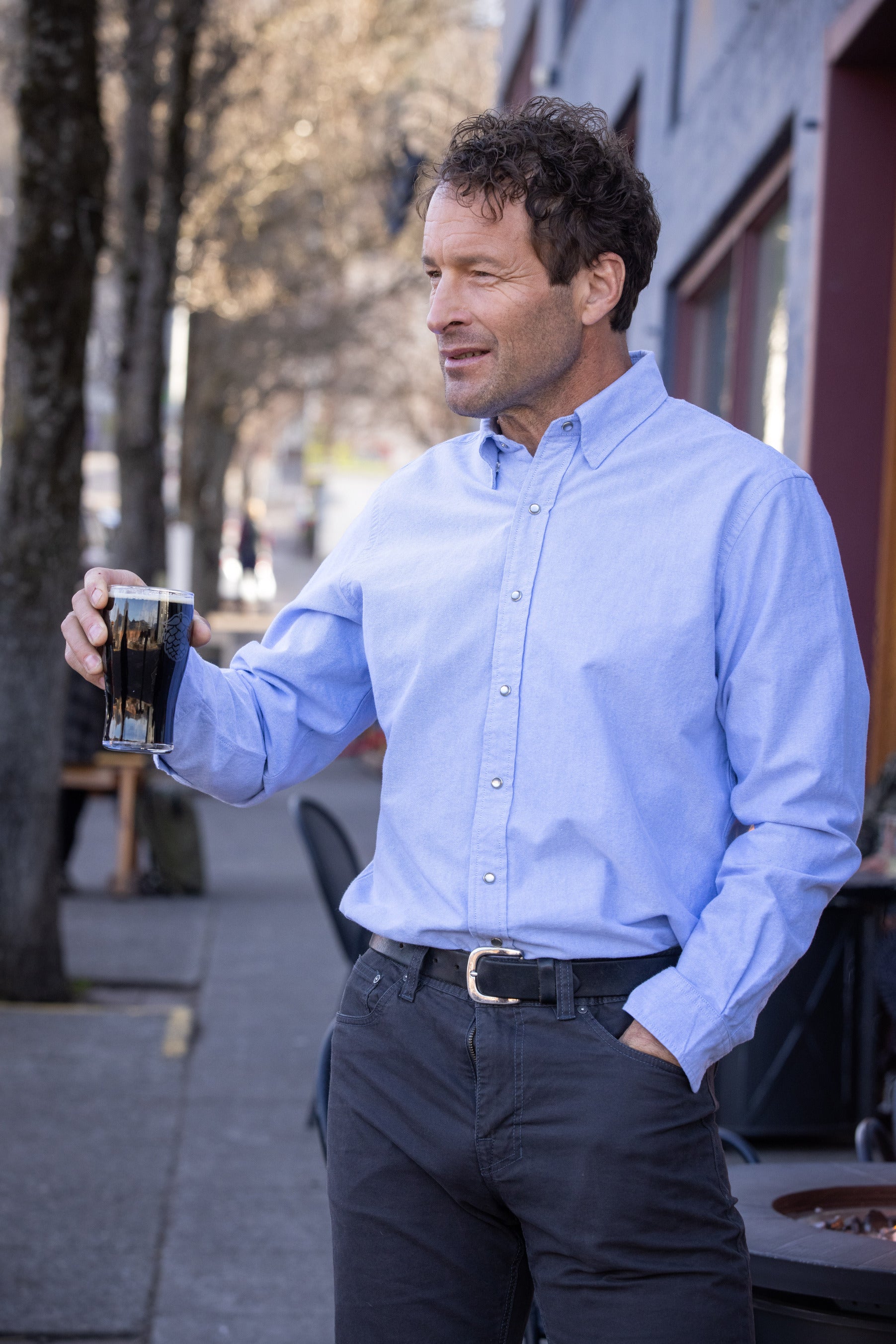 A man in a light blue Unbuttoned shirt holding a drink on a city sidewalk